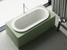 Oval built-in bathtub with green tile surround, freestanding faucet, and curtain by the window.